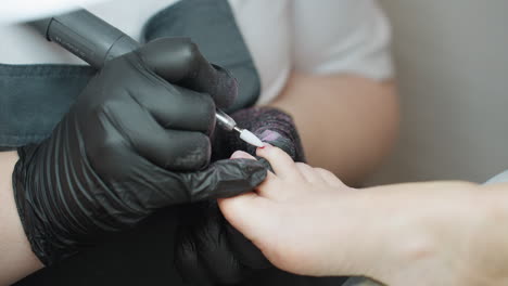 close up view of nail technician wearing black gloves using electric file on client toe during pedicure procedure, with focus on precision and nail care in professional salon environment