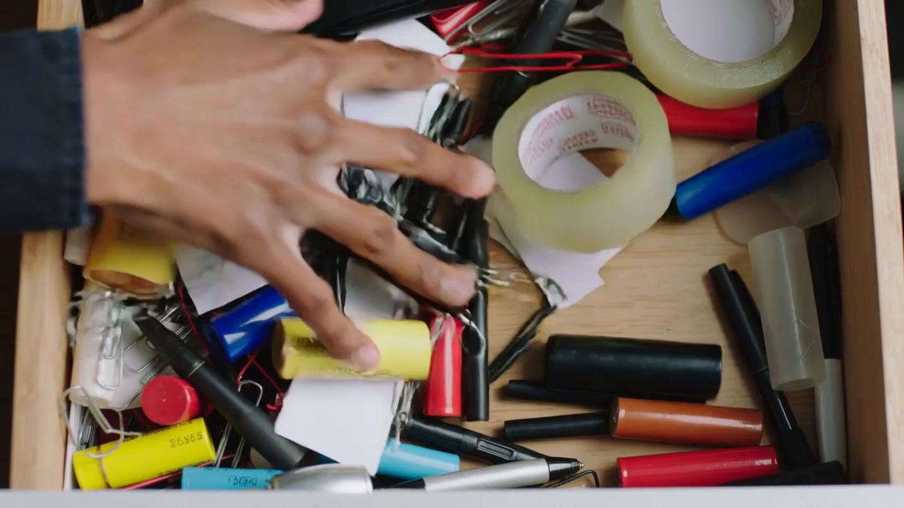 Reaching right hand in dark shirt searching shallow wooden desk drawer, moving tape and batteries
