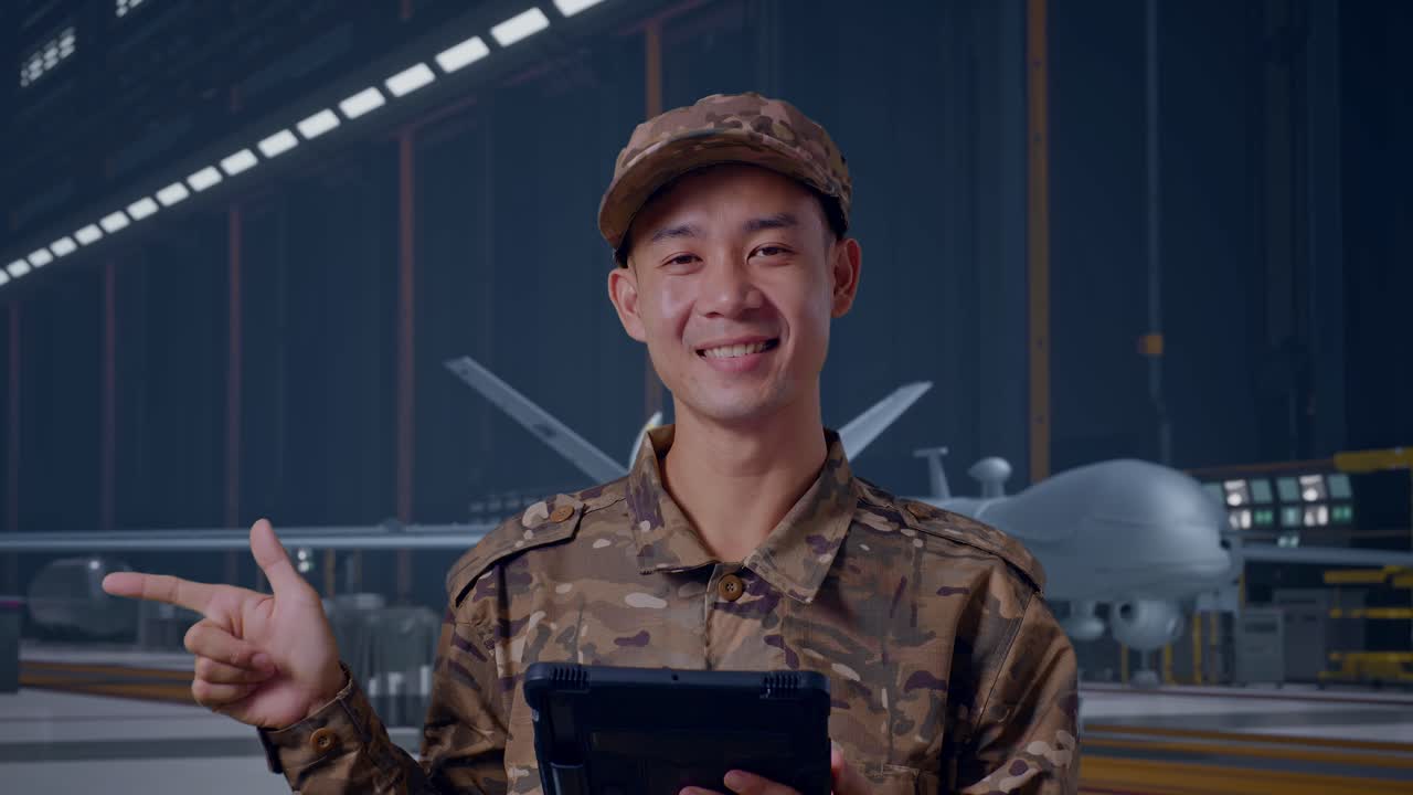 Prajurit Pria Asia Tampak Dekat Menggunakan Tablet dan Menunjuk ke Samping Sambil Berdiri dengan Drone Militer Modern di Hangar Pemeliharaan