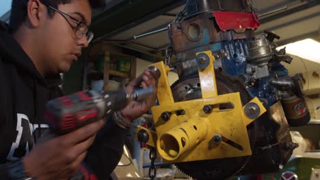 Joven-Mecánico-Desatornillando-Tornillos-Con-Un-Taladro-Del-Soporte-Del-Motor