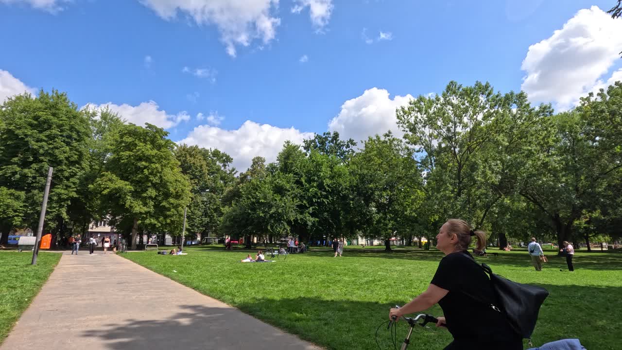 A person rides a bicycle along a paved path in a vibrant Berlin park on a sunny day, with lush greenery and scattered visitors. Camera pans smoothly