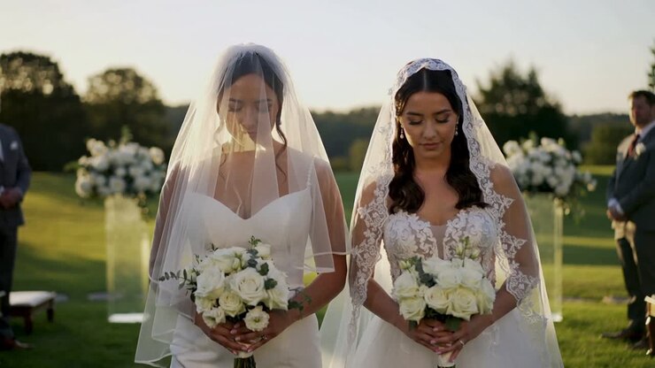 Lowering heads after officiant cue, brides in gowns wearing veils on grassy lawn, holding bouquets