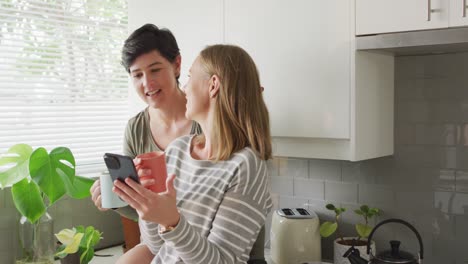 Kaukasisches-Lesbisches-Paar-Hält-Kaffeetassen-Und-Umarmt-Sich-In-Der-Küche-Zu-Hause