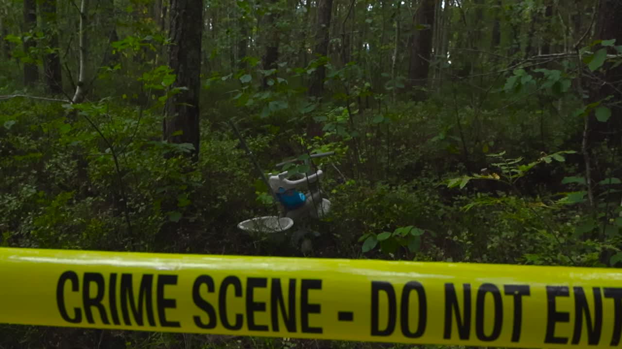 Police forensic yellow colored crime scene tape ribbon in front of a dark and lush day time forest area where a abandoned child or kids bicycle is in the moss in between large trees during cloudy day