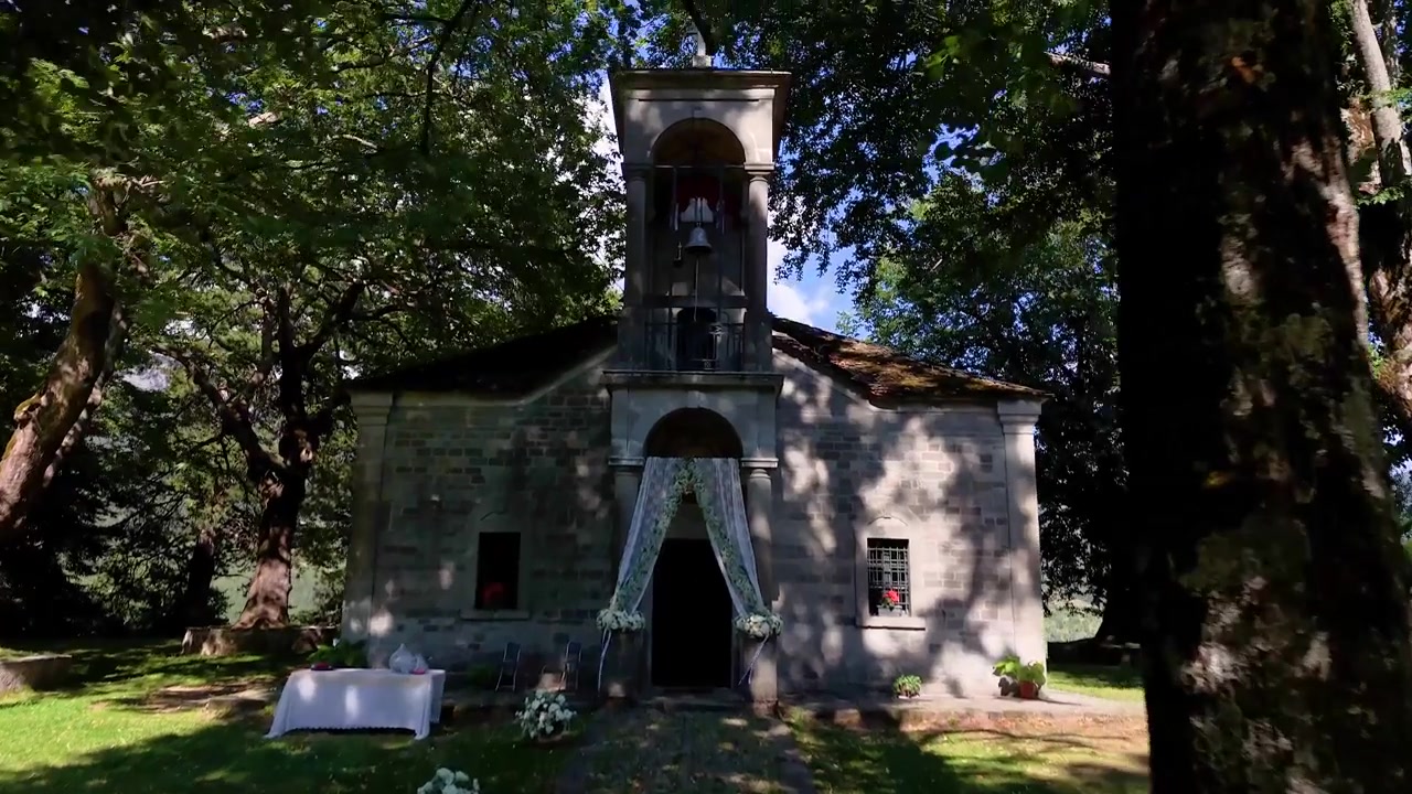 Una piccola cappella in pietra si erge tra alberi alti, il suo ingresso adornato con decorazioni floreali nuziali bianche alla brillante luce di mezzogiorno