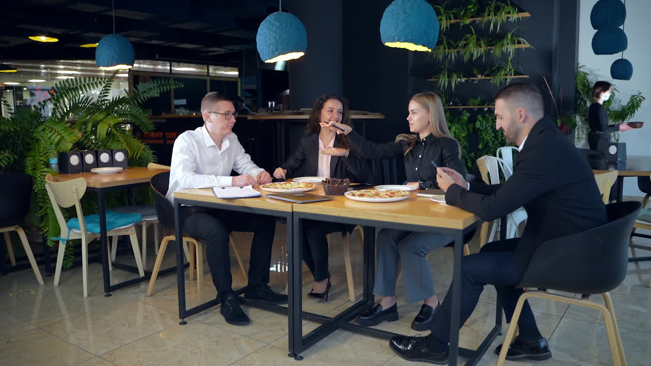 Teammates sitting in a buffet at lunchtime. Man and two women speaking and gesturing. Other man is concentrated on his smartphone.