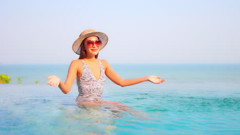 A-playful,-young,-fit-woman-in-a-zebra-print-bathing-suit-splashes-water-into-the-air-as-she-sits-on-the-edge-of-an-infinity-edge-pool-with-an-ocean-background