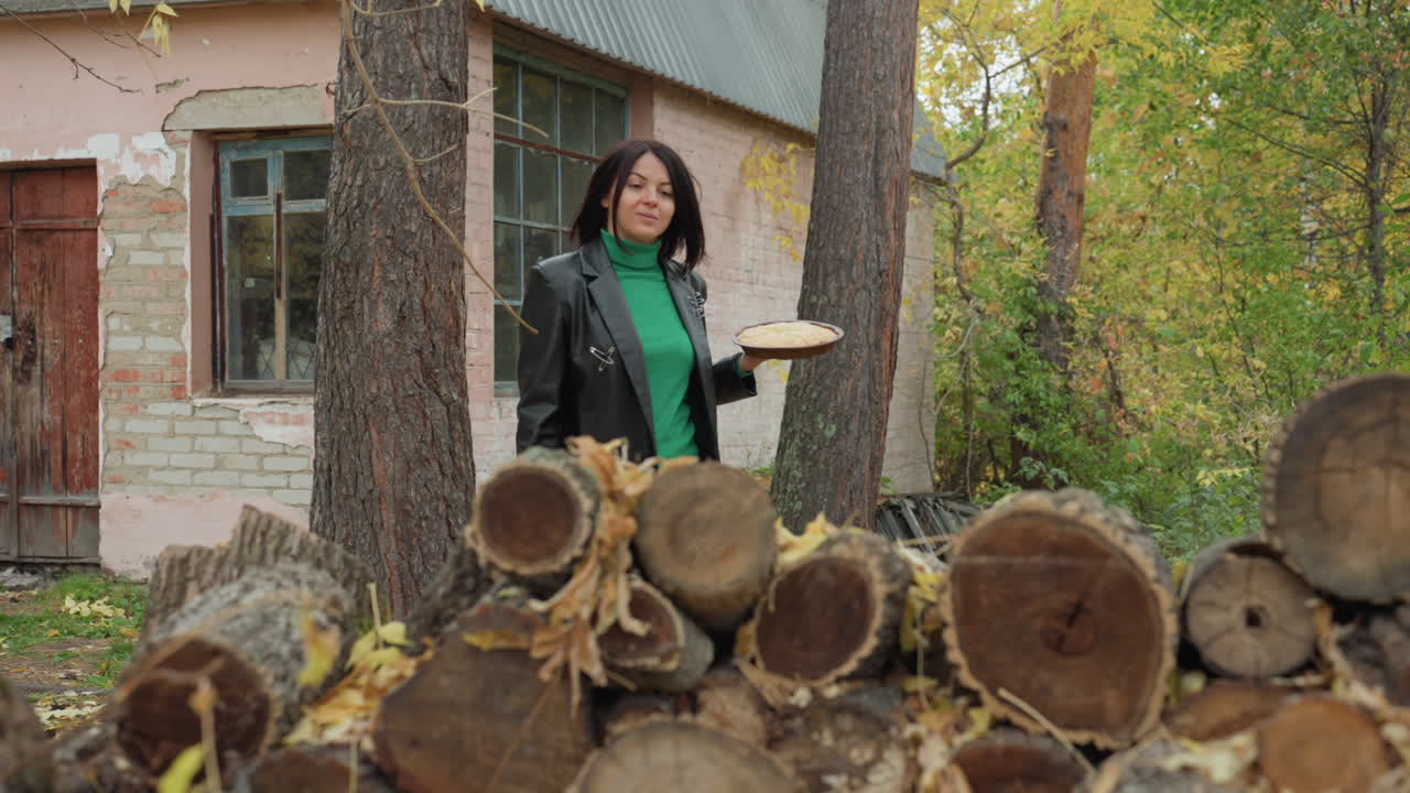 woman in leather jacket near logs outside pink cottage. wears green sweater and black coat, carrying wooden plate while walking among stacked firewood and tall pine trees. autumn leaves carpet ground
