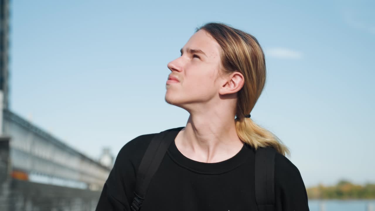 Young individual looks sideways. Youthful person glances to side. Young man with backpack gazes sideways attentively. Youthful individual carrying backpack observes objects passing by on side