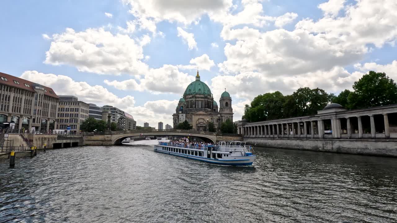 A river cruise boat glides past Berlin Cathedral and museum buildings on the Spree River under bright daylight, captured in a wide, steady shot