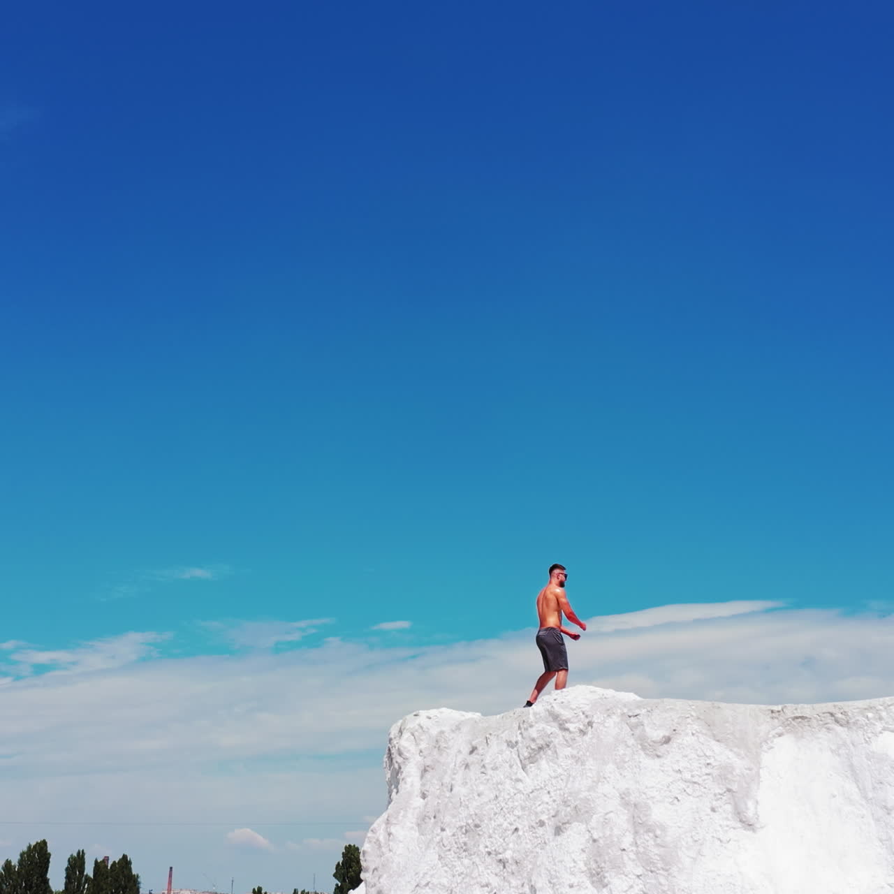 Sportive man in shorts showing his trained body on white hill. Back view of a strong athlete standing in pose on the mountain under blue sky in summer.