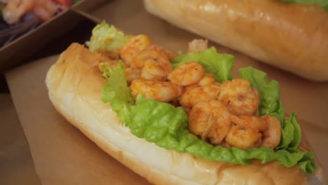 close-up of a grilled shrimp sandwich layered with crisp lettuce in a soft bun, surrounded by other plated seafood items in warm indoor lighting.
