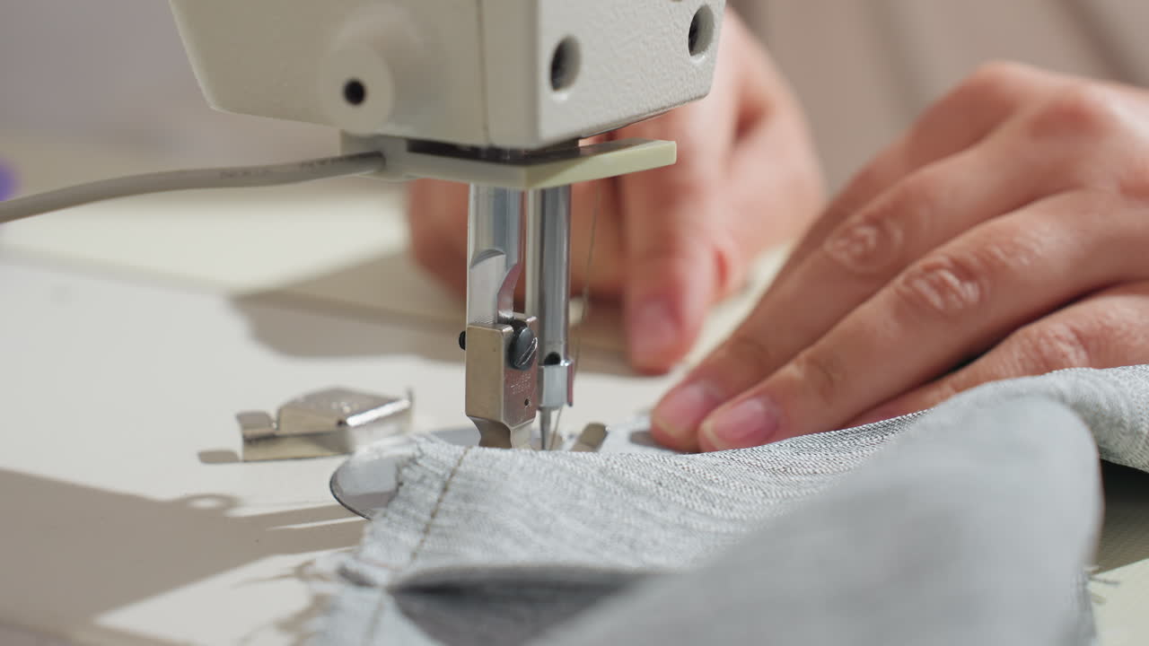 Close up view of sewing machine needle stitching gray fabric as female hand gently guides material under presser foot during garment production process in brightly lit tailoring workspace