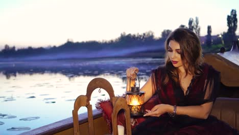 pretty woman in dress sitting in a boat with candelabrum in the evening. beautiful lady holding lamp in hands at sunset over the natural background