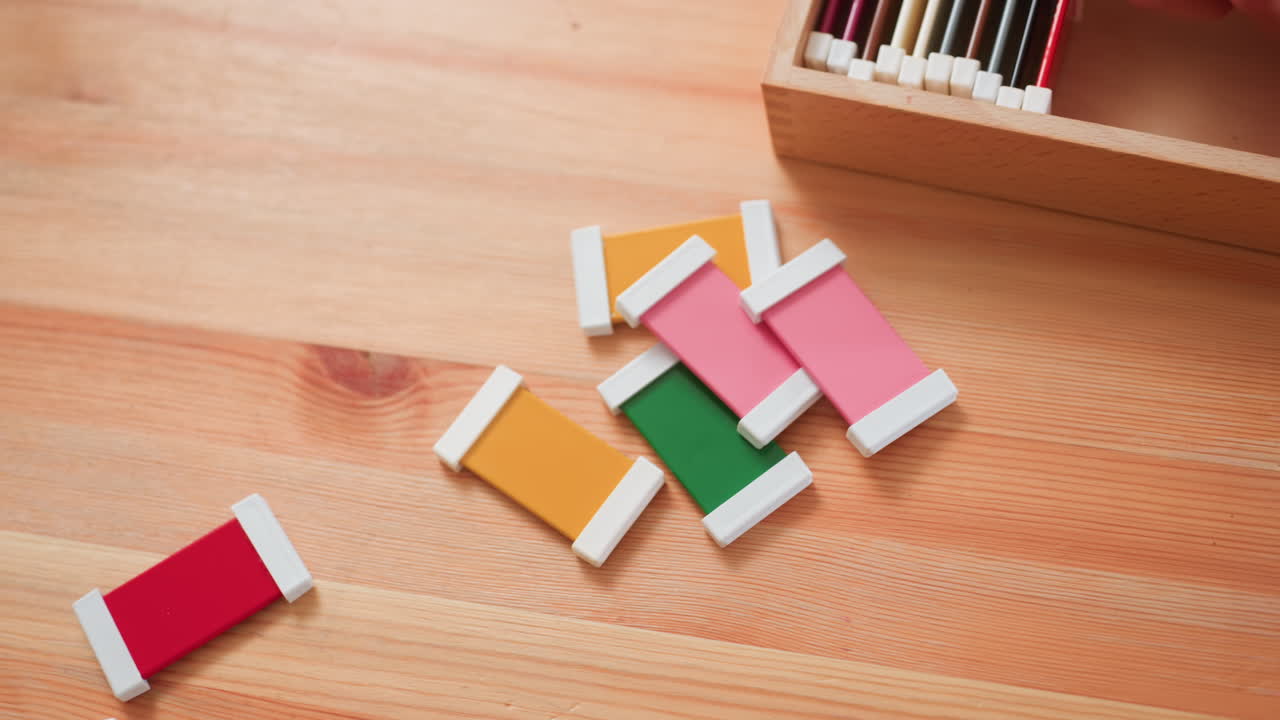 Adult arranges colorful blocks back into wooden box on table, hand placing pieces neatly, pink, green, yellow, red visible, educational material cleanup, order and guidance