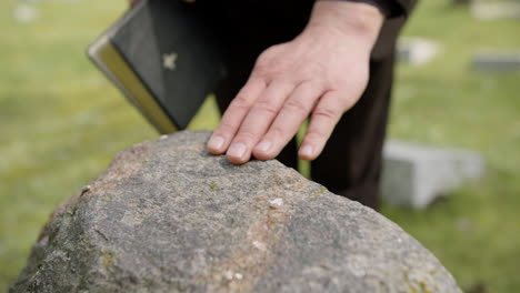 close up view of man hand touching a tombstone while holding a bible in a graveyard 2