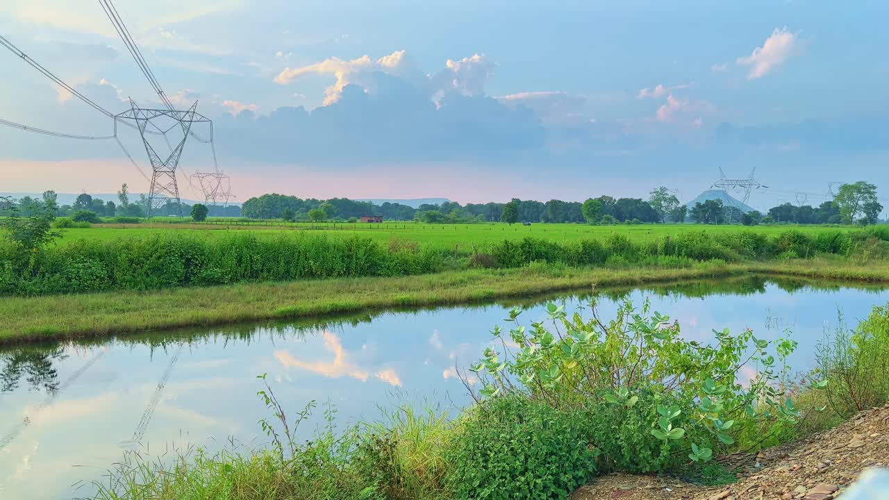 Static shot of towering power lines crossing a green farmland with their reflection in still water, blending technology and nature in a peaceful rural setting under a soft blue sky