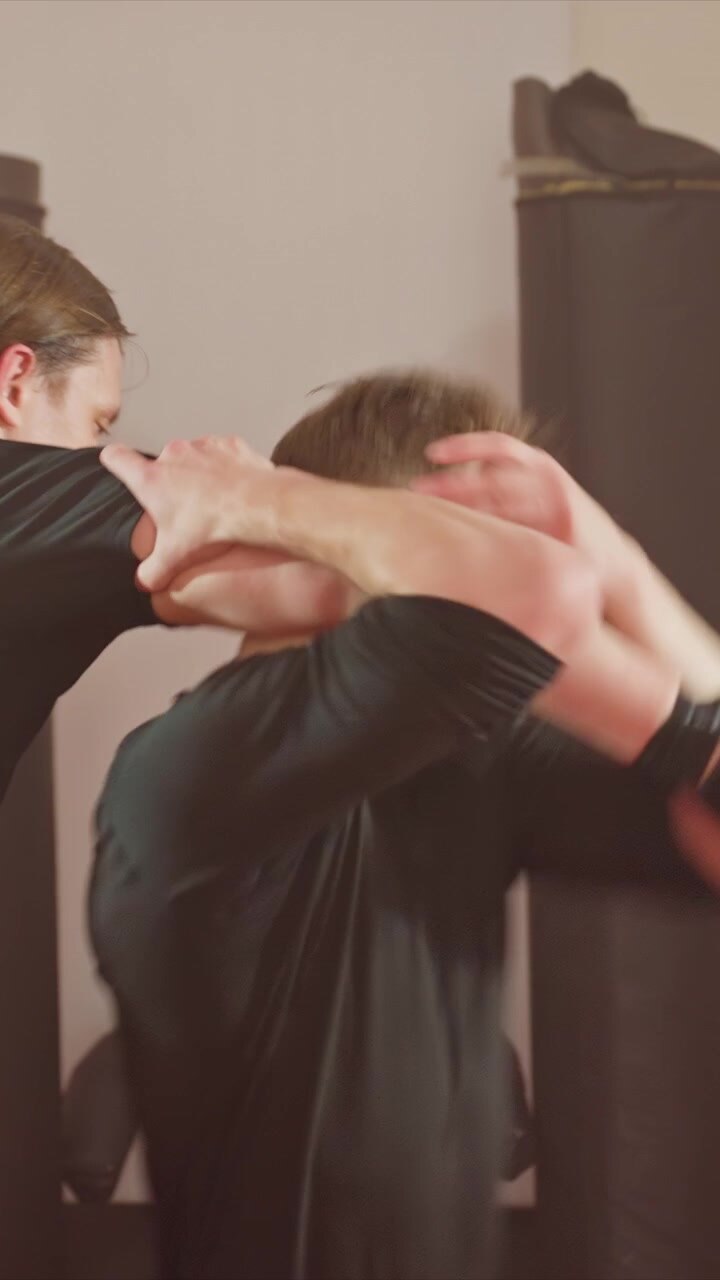 Two young men engage in martial arts training with one applying pressure on other who is lying. Scene takes place indoors against plain wall. Close up shots focus on upper body