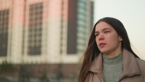 a close-up front shot of a young girl wearing a peach jacket and grey inner sweater. her mouth is slightly open, and her expression appears pensive, with the backdrop of tall buildings