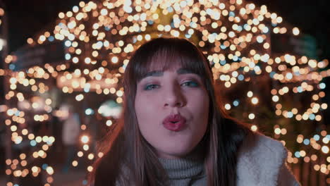 joven hermosa con labios rojos soplando un aire frío de invierno a la cámara y sonriendo con las luces naranjas parpadeantes en el fondo en el mercado de navidad en cámara lenta