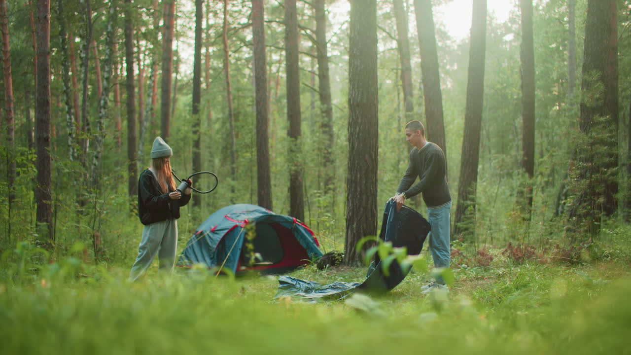 Young man lifts tent fabric while spraying it in forest clearing as woman watches nearby holding hose, surrounded by trees and camping gear