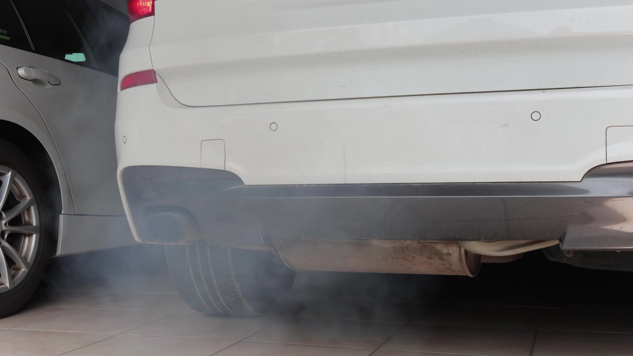 Beyaz bir aracın arka tamponunun yakın çekim sabit görüntüsü. Motor çalıştırıldığında egzoz borusundan kalın gri egzoz dumanı çıkıyor