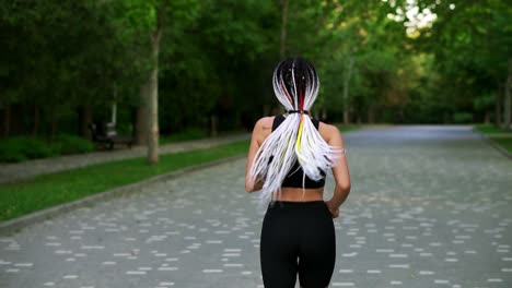 Rear-view-of-european-woman-with-black-and-white-dreadlocks-running-by-local,-green-park-in-the-city.-She-is-exercising-for-good
