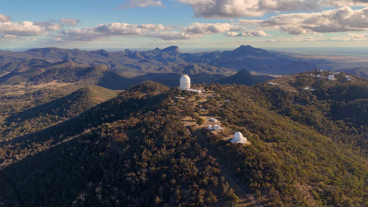 Aerial drone footage glides over a forested mountain ridge, revealing a large observatory dome and telescopes under warm, late afternoon sunlight