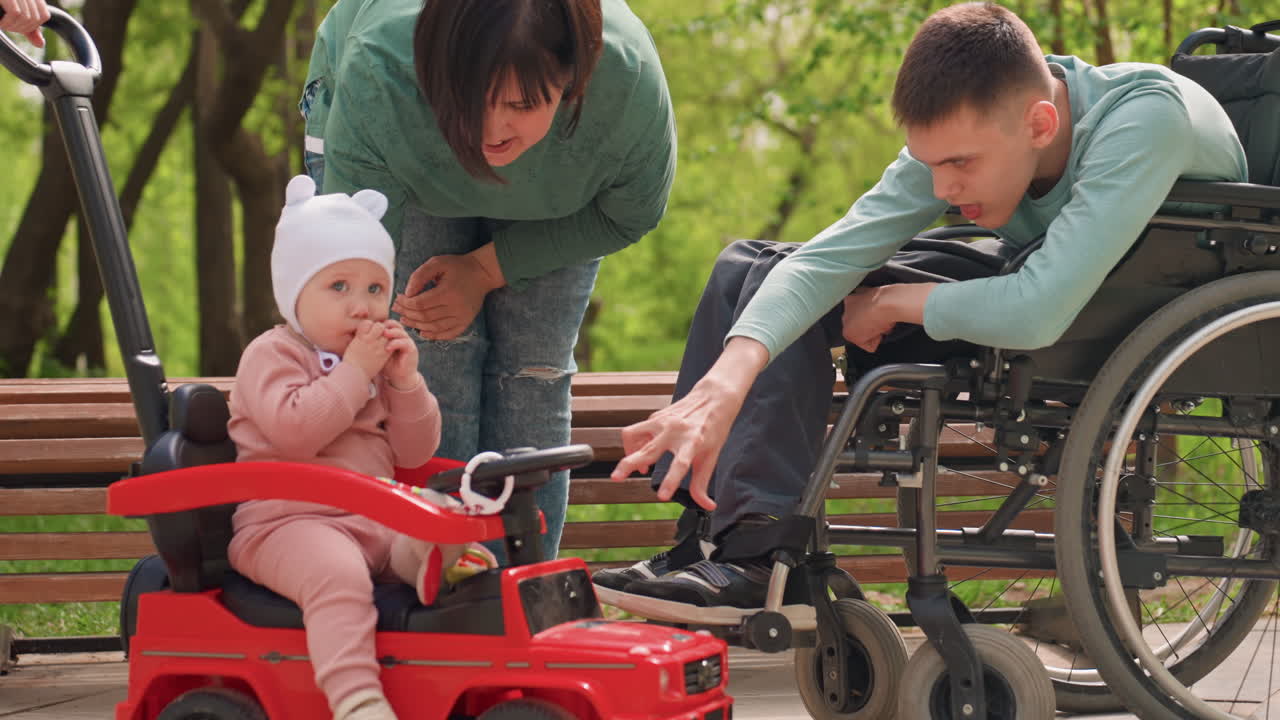 Young Man In Wheelchair Reaches Toward Toddler Seated In Red Ride Car, Gentle Interaction And Curiosity In Park Setting, Caregiver Nearby And Warm Inclusive Family Scene With Sunny Greenery