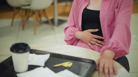 lower view of woman wearing pink dress gently rubbing her stomach with right hand, while her left hand rests on the table, a tray with a coffee cup and napkins lies in front of her