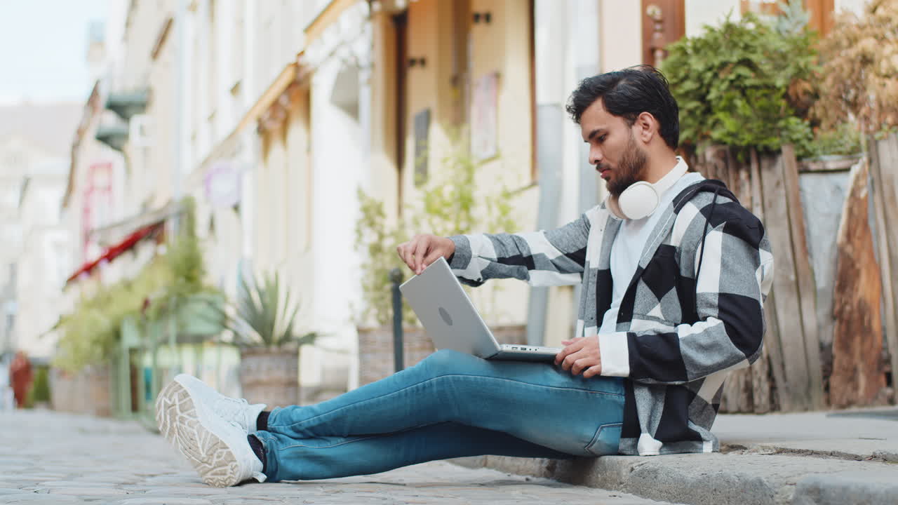 Indian man freelancer sitting on street closes laptop screen finishing work smiling in city outdoors