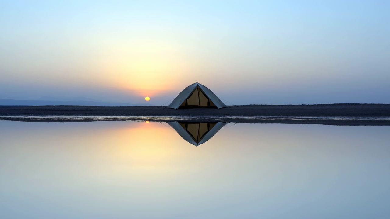 Static wide view shows solitary tent centered on dark shore while calm water mirrors its shape and warm low sunlight, with hazy hills lining distant horizon under blue-orange sky.
