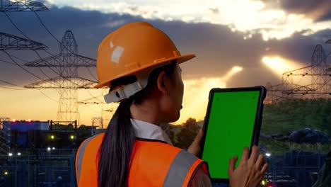 vista posterior cercana de una ingeniera asiática con casco de seguridad trabajando en una tableta de pantalla verde y mirando a su alrededor cerca de la torre de alto voltaje