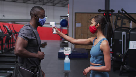 Mujer-Caucásica-En-Forma-Con-Máscara-Facial-Midiendo-La-Temperatura-De-Un-Hombre-Afroamericano-En-Forma-En-El-Gimnasio