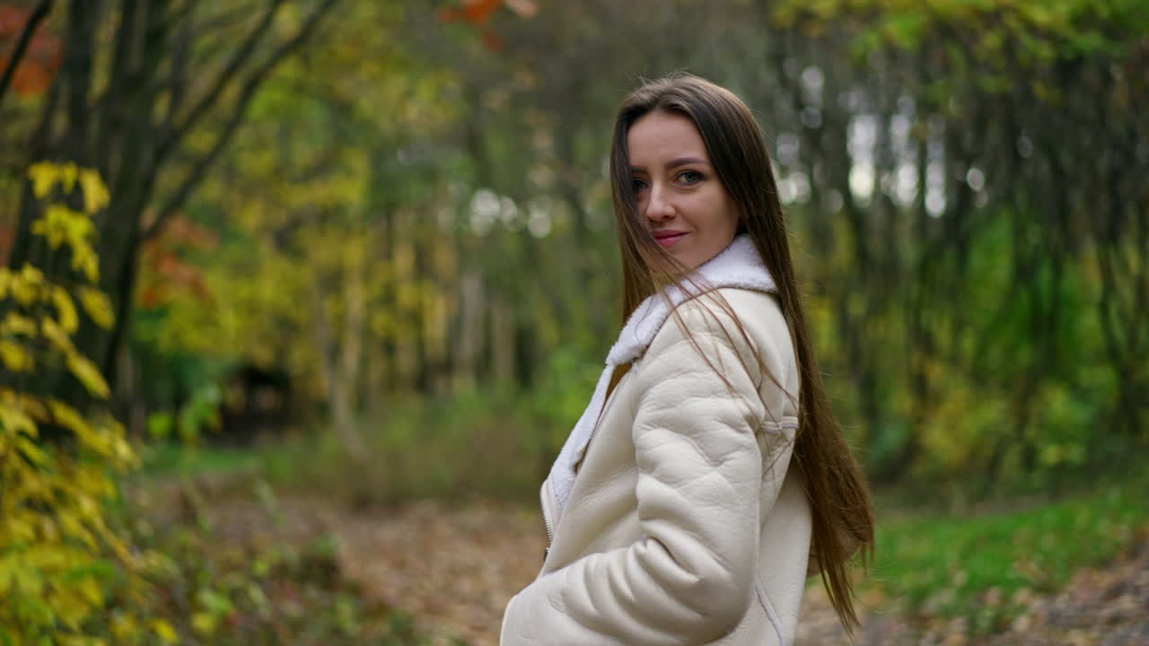 Long-haired brunette woman standing in the autumn park turns around to the camera. Lady looks into camera slowly approaching to her face. Blurred backdrop.