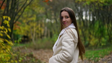 long-haired brunette woman standing in the autumn park turns around to the camera. lady looks into camera slowly approaching to her face. blurred backdrop.