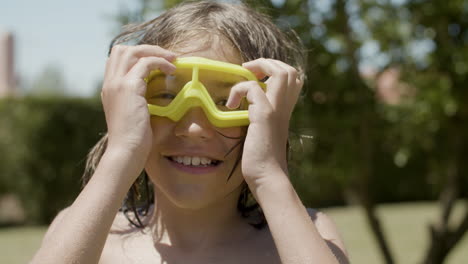 vista frontal de un niño feliz con el torso desnudo y gafas para deportes acuáticos mirando la cámara en el jardín de casa