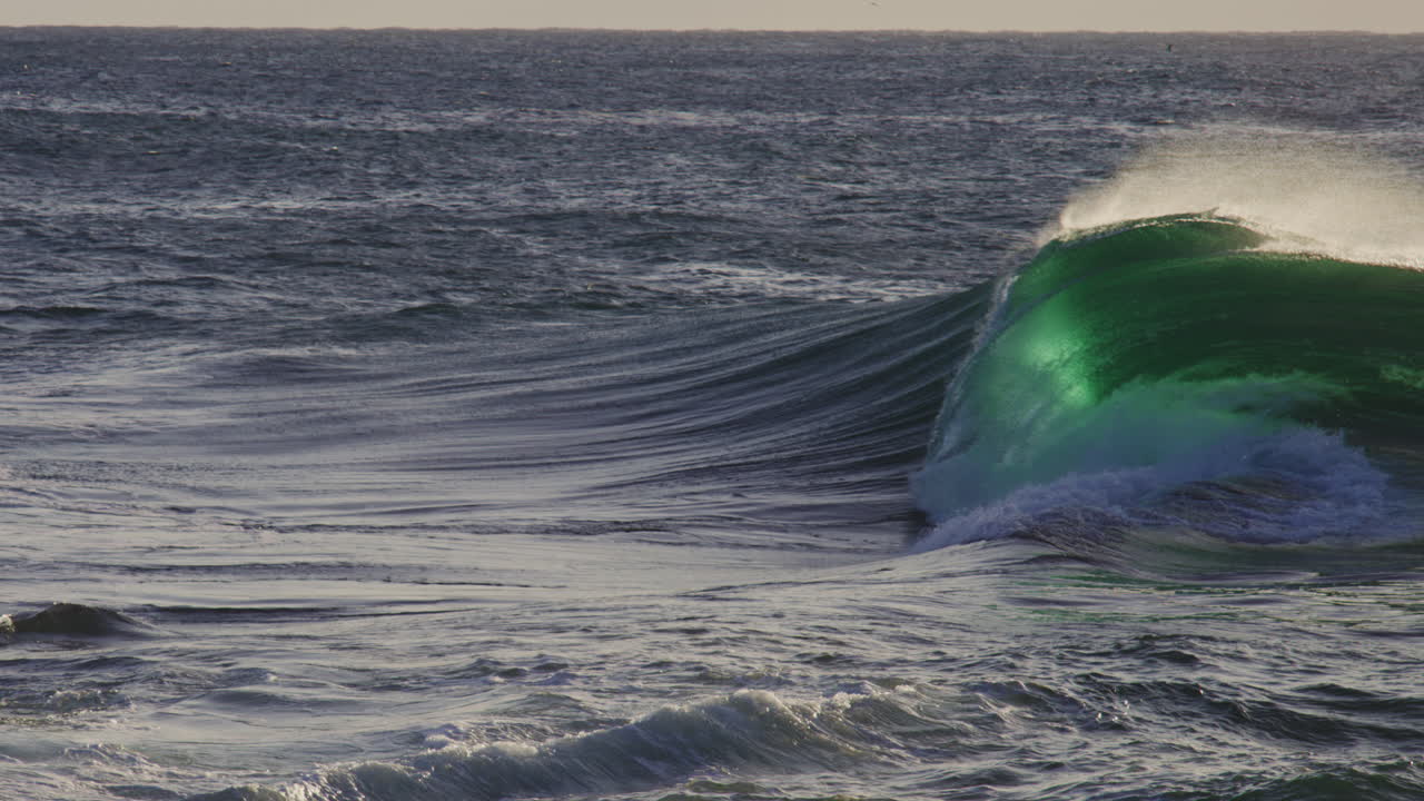 Super slow motion wave rises glowing green with mist and ocean spray