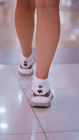 vista de la pierna de la dama en zapatillas blancas caminando por el piso de baldosas pulidas con reflejos de luces superiores que crean un brillo suave, sombras de personas y luces vibrantes en el piso brillante