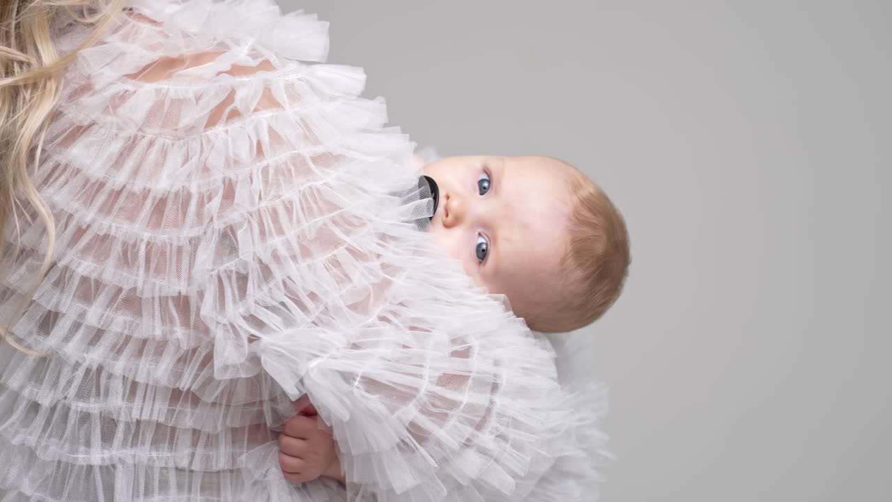 Unrecognized long-haired blond woman holding a baby. Tiny infant with pacifier looking at camera. White backdrop.