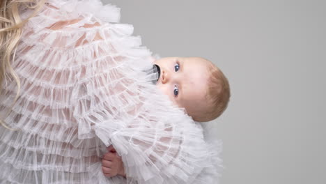 unrecognized long-haired blond woman holding a baby. tiny infant with pacifier looking at camera. white backdrop.