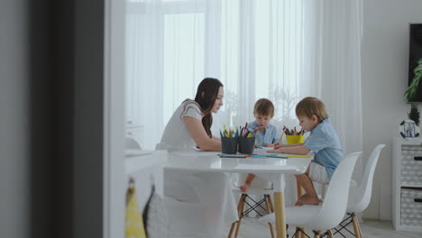 Mom-helps-sons-learn-to-draw-doing-homework-preschool-preparation-at-home-sitting-in-the-white-kitchen.-Two-children-brothers-draw-a-portrait-of-mother-together