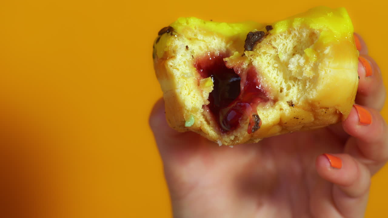 A Hand holds a yellow-glazed Berliner donut with a large bite revealing its vibrant cherry jam filling, against an orange background.