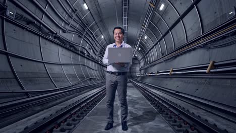 full body of an asian male professional worker standing with his laptop in underground subway tunnel, he is looking at the camera with a smile
