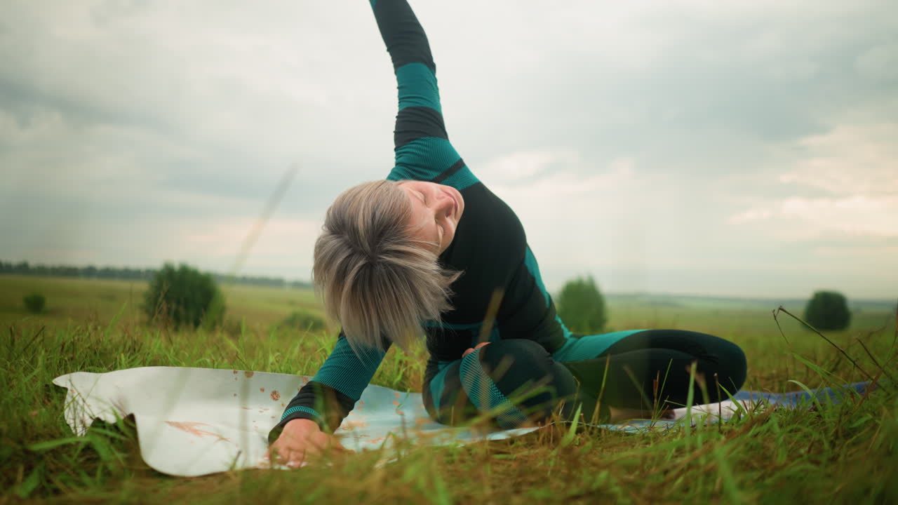 mujer avanzada acostada de un lado en una alfombra de yoga practicando la postura de flexión lateral con el brazo extendido en un vasto campo de hierba bajo un cielo nublado, rodeada de naturaleza y árboles en la distancia