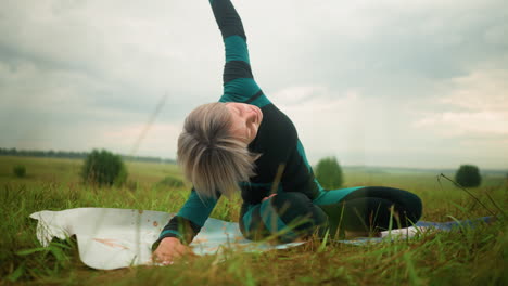 advanced woman lying on one side on yoga mat practicing side bend pose with arm extended in a vast grassy field under cloudy skies, surrounded by nature and trees in the distance