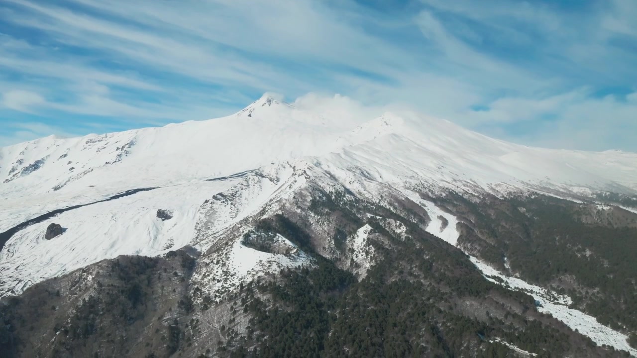Le creste montuose innevate del vulcano Etna si ergono sopra pendii boscosi scuri sotto un cielo azzurro.