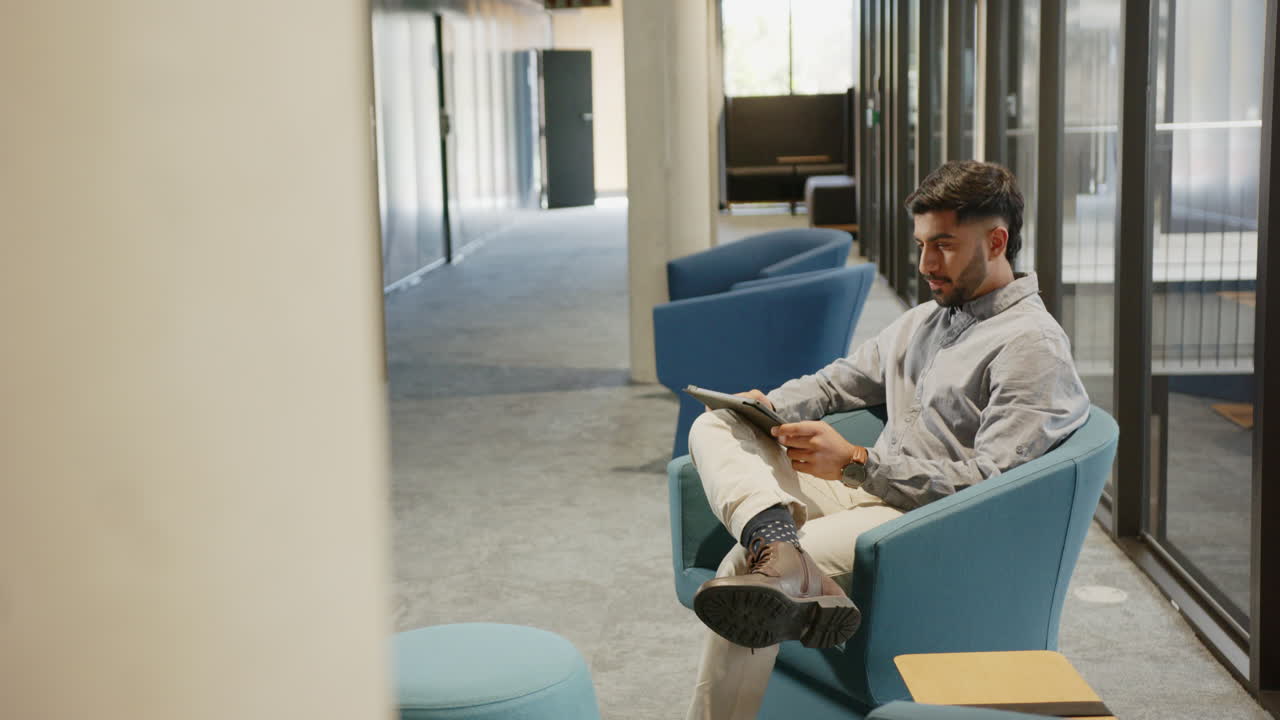 Reading tablet, Indian man sitting in modern office lounge area, relaxing