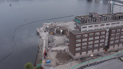 drone shot orbiting building revealing building with three diggers working on removing rubble by the water in liljeholmsviken, liljeholmen, stockholm, sweden