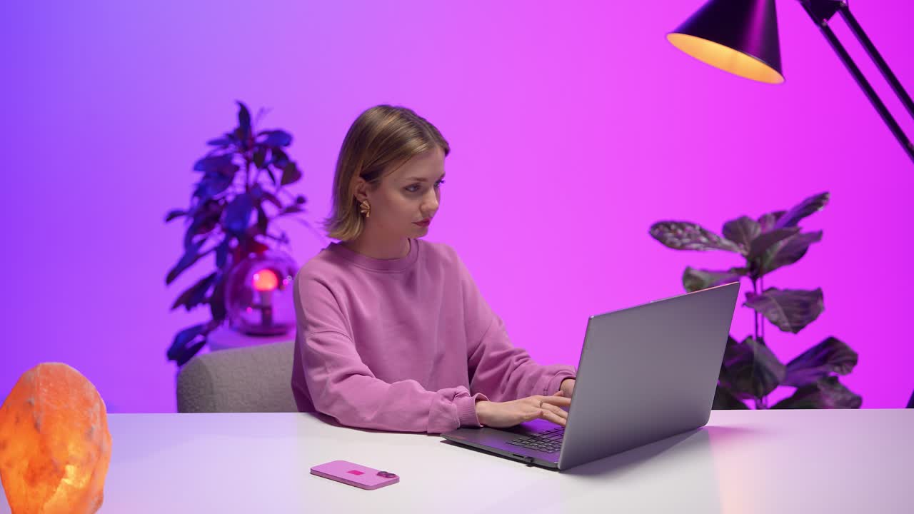 A woman in a pink sweatshirt uses a smartphone while working on a laptop at a desk. Surrounded by plants, purple lighting and soft decor, the scene reflects a balanced, tech-driven modern workflow